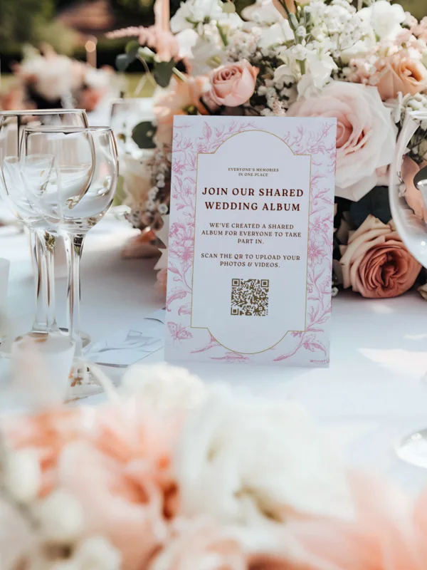 Pink floral QR card standing among roses and wine glasses on a set reception table
