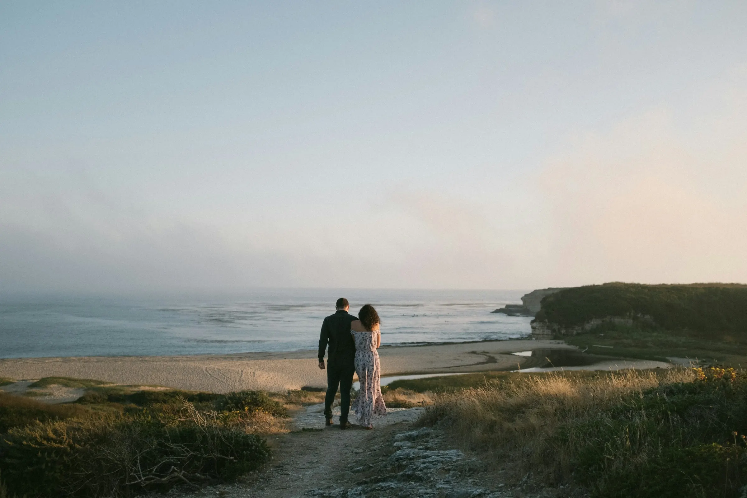 Una pareja de espaldas paseando por un sendero costero de hierba hacia el mar bajo un suave cielo vespertino