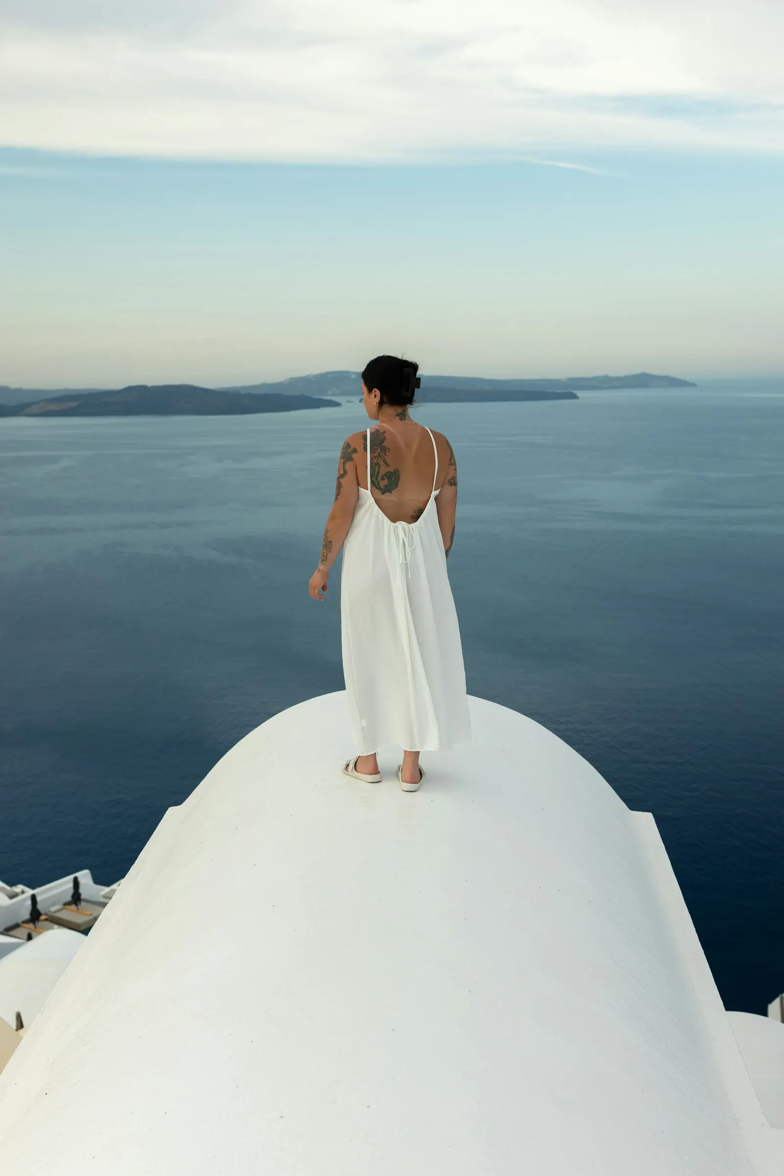 Una novia de espaldas con un sencillo vestido lencero blanco en una terraza encalada de Santorini, contemplando el mar Egeo
